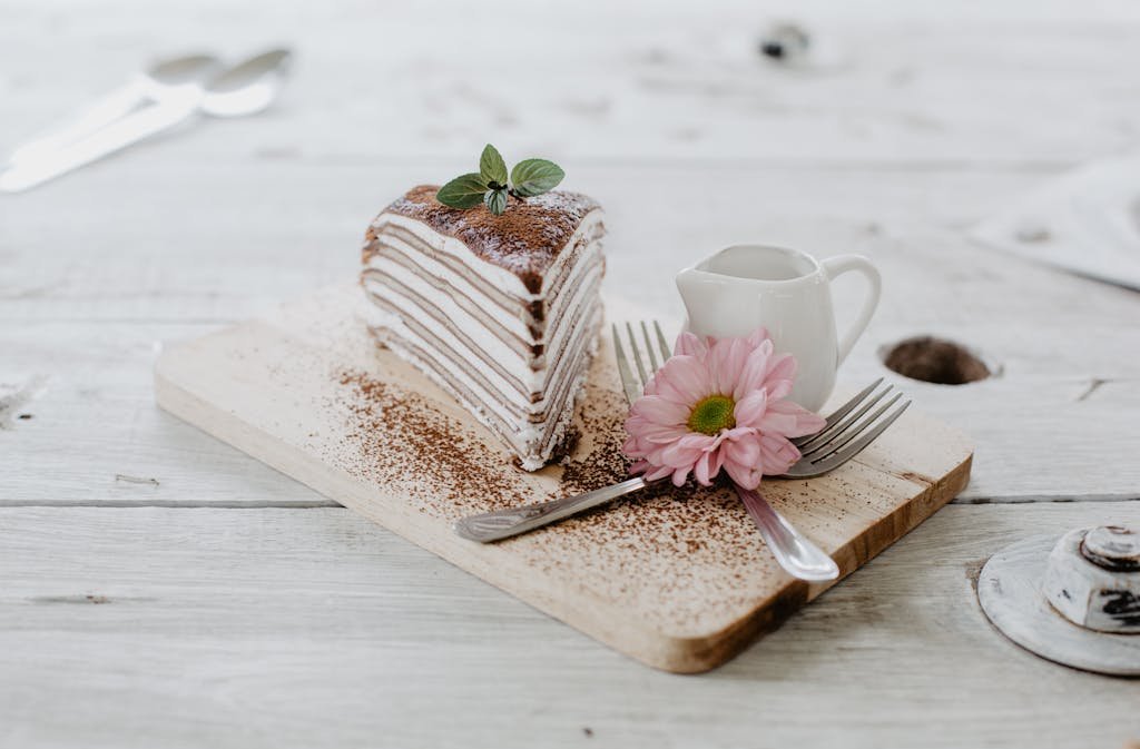 From above of appetizing piece of cake decorated chocolate powder and mint leaves served near ceramic creamer and forks with light pink chrysanthemum on top placed on wooden board