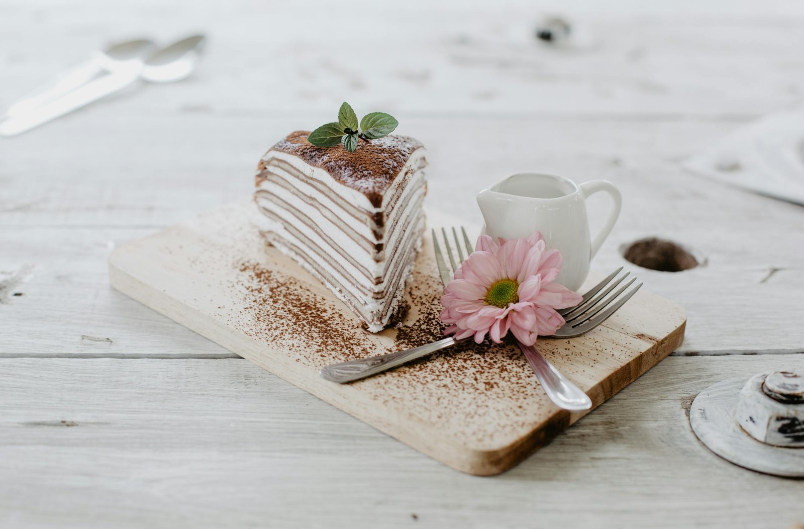 From above of appetizing piece of cake decorated chocolate powder and mint leaves served near ceramic creamer and forks with light pink chrysanthemum on top placed on wooden board