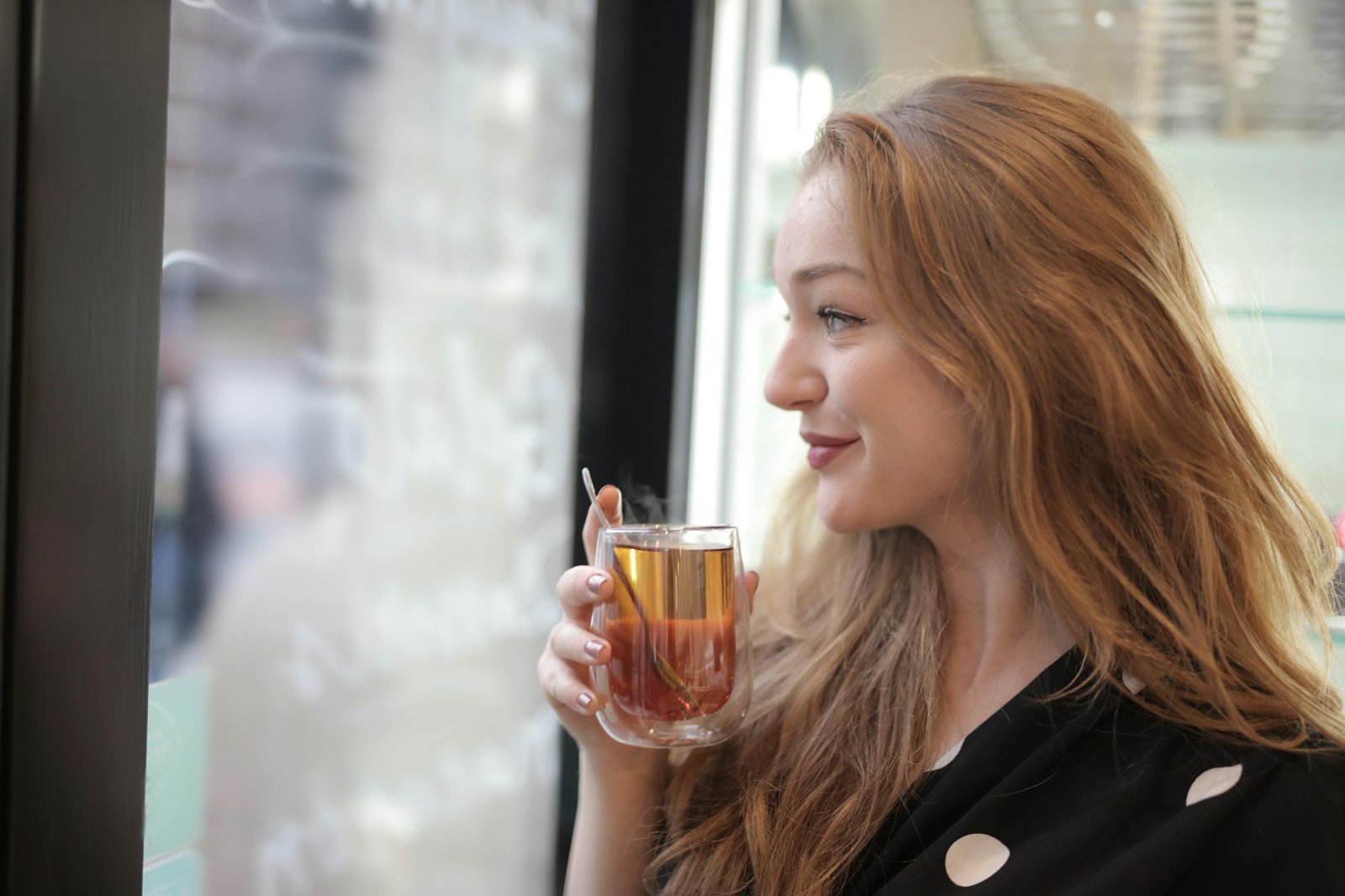 Side view of joyful female in polka dot dress standing with glass cup of hot beverage and looking out of window while relaxing in cafeteria during weekend